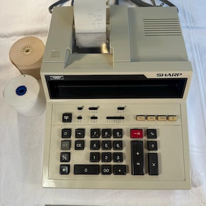 May include: A vintage Sharp Comet calculator with a built-in printer. The calculator is beige with black keys and a paper roll. A measuring tape shows 10 inches. The calculator prints a receipt.