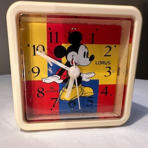 May include: Vintage Lorus quartz alarm clock featuring Mickey Mouse. The clock has a cream-colored square frame and a colorful face with Mickey Mouse, red, yellow, and blue sections, and white hands. The clock face displays the numbers 1 through 12.