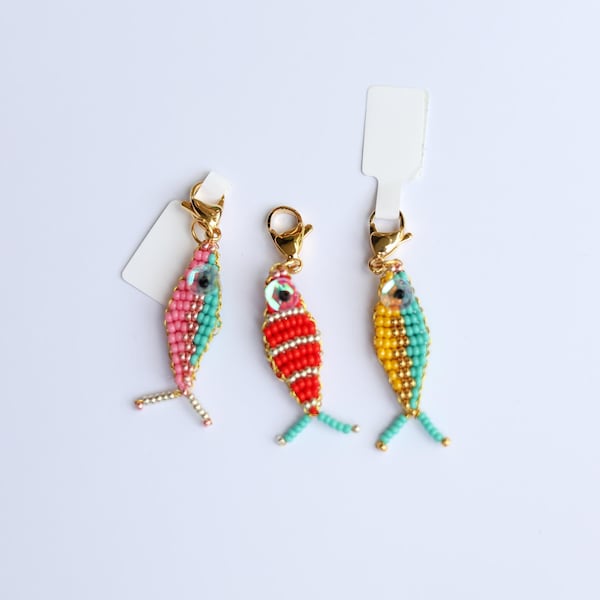 以下が含まれることがあります： ピンク、赤、黄色のビーズで作られた魚のチャームが3つ。ゴールドカラーの留め金が付いています。それぞれの魚は異なる配色で、ターコイズとゴールドのアクセントがあります。白い背景に配置されています。