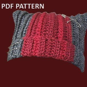 May include: A handmade crochet hat pattern in red and gray, secured with safety pins. The text "PDF PATTERN" is displayed at the top. The hat is made of thick yarn, with a ribbed texture. The color scheme is a mix of red and gray.