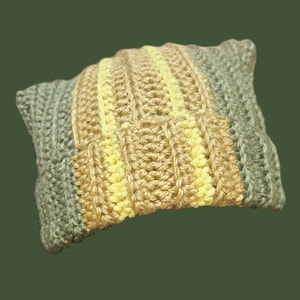 May include: A rectangular crochet pillow featuring vertical stripes in alternating colors of sage green, tan, and pale yellow. The textile is constructed with a textured, ribbed stitch pattern that runs horizontally across the entire surface.
