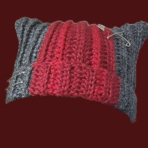 May include: A rectangular crochet beanie featuring a ribbed texture with a central vertical panel in deep red and side panels in charcoal gray. The hat is accented with three silver-toned metal safety pins attached to the fabric surface.