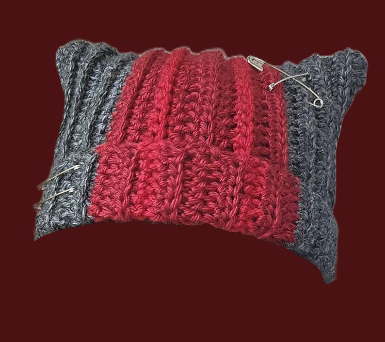 May include: A rectangular crochet beanie featuring a ribbed texture with a central vertical panel in deep red and side panels in charcoal gray. The hat is accented with three silver-toned metal safety pins attached to the fabric surface.