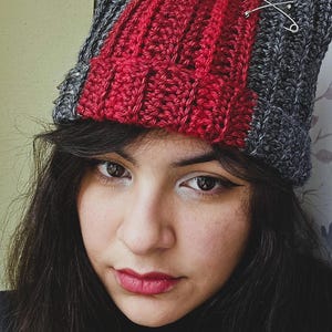 May include: A crocheted cat-ear beanie featuring a color-blocked design with dark gray sides and a wide, vertical red stripe down the center. The hat is embellished with silver-toned safety pins attached to the top and side sections.