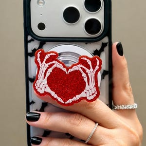May include: A smartphone case featuring a black and white bat pattern, equipped with a circular phone grip. The grip displays a red glitter heart shape formed by two white skeleton hands, creating a distinct contrast against the sparkling red background.