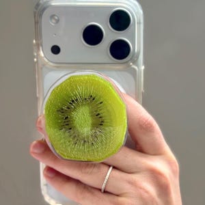 May include: A circular phone grip featuring a realistic, high-resolution print of a sliced kiwi fruit with a vibrant green center, small black seeds, and a translucent edge, attached to the back of a clear smartphone case with a triple-lens camera.