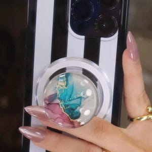 May include: A black and white striped phone case with a pop socket. The pop socket has a clear base and a colorful design with blue, pink, and gold accents. The phone case is held by a person.
