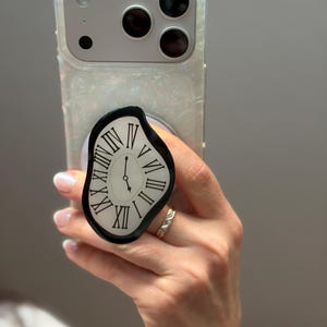 May include: A clear phone case with a pearlescent shimmer holds a smartphone. A black and white pop-socket with a melting clock design is attached. The clock face has Roman numerals and black hands, with the time showing approximately 5 o'clock.