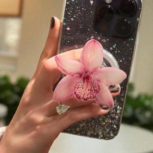 May include: A black phone case with a glitter and star design. A pink orchid flower is held in front of the phone. A silver ring with a pear-shaped diamond is on the person's finger.