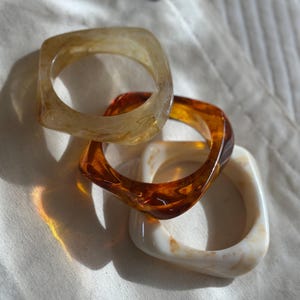 May include: Three square-shaped resin bangles in shades of amber, brown, and cream. The bangles are stacked on top of each other, with the light reflecting off their glossy surfaces. The jewelry is set against a white fabric background.