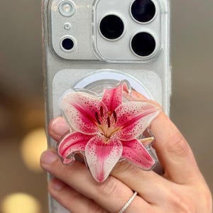 May include: A clear, glittery smartphone case featuring a circular phone grip attachment. The grip is decorated with a detailed, realistic illustration of a pink and white lily flower with dark pink speckles and prominent dark brown stamens.