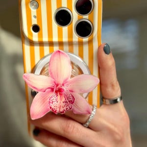 May include: A phone case with a vertical orange and white striped pattern. A pink orchid flower is attached to the back of the phone case. The phone case has a clear ring holder.