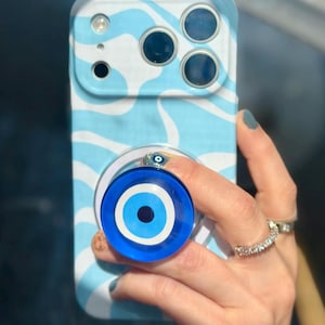 May include: A light blue and white patterned phone case with a matching pop socket featuring a blue eye design. The phone case has a wave-like pattern and a camera cutout.