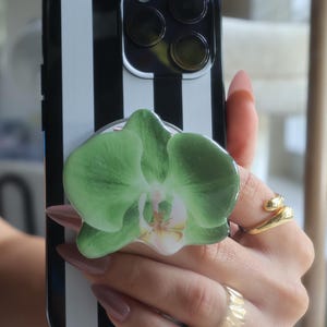 May include: Black and white striped phone case with a green orchid flower phone grip. The phone case has a camera bump with three lenses. The phone grip is a light green color with a white and yellow center.