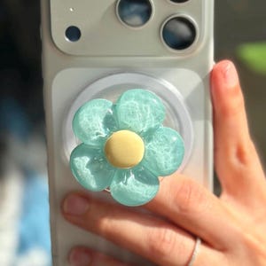 May include: A light blue, translucent, flower-shaped phone grip attached to the back of a white smartphone. The flower features five rounded petals with a smooth, solid pale yellow circular center, creating a glossy, textured appearance.