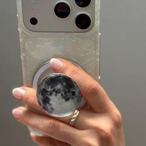 May include: A clear phone case with a pearlescent sheen, holding a smartphone with a triple-lens camera. A round phone grip featuring a detailed moon design is attached. The grip is black and white, and the phone is held by a hand with a gold ring.