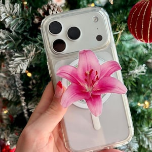 May include: Clear phone case with a pink lily flower design. The phone case has a white magnetic ring and is held in front of a Christmas tree.