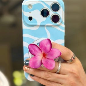May include: A light blue and white patterned phone case with a pink flower phone grip. The phone case has a wave design. The flower grip is a vibrant pink with a red center. The phone case is held in a hand.