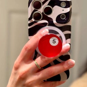 May include: A phone case with a black and white zebra stripe pattern. A red, round phone grip with the number 5 is attached. The phone case is held by a hand with a gold ring.