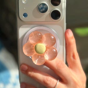 May include: A light-colored smartphone featuring a circular, collapsible phone grip attached to the back. The grip is shaped like a flower with translucent, peach-colored petals and a solid, pale yellow circular center, providing a secure hold.