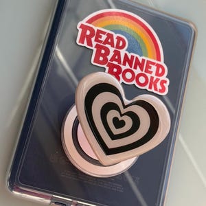 May include: A dark blue electronic device in a clear case featuring a rainbow sticker with the text READ BANNED BOOKS and a heart-shaped phone grip with a black and white concentric heart pattern attached to the back of the device.