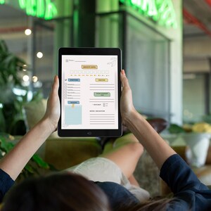 May include: A black tablet displaying a wellness tracker with sections for anxiety level, symptoms, and emotions. The tablet is held by a person, with a yellow headband visible in the foreground. The background features green neon lights.