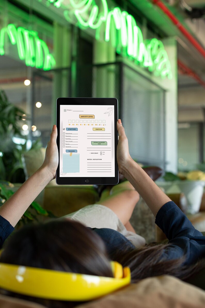 May include: A black tablet displaying a wellness tracker with sections for anxiety level, symptoms, and emotions. The tablet is held by a person, with a yellow headband visible in the foreground. The background features green neon lights.