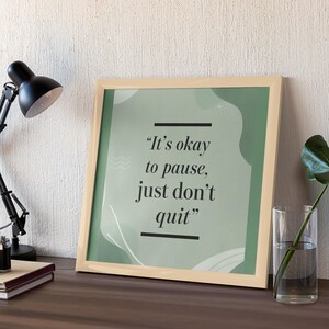 May include: A framed print with the quote "It's okay to pause, just don't quit" in black text on a green background. The frame is light beige, and the print is displayed on a wooden surface with a black desk lamp and a glass vase.