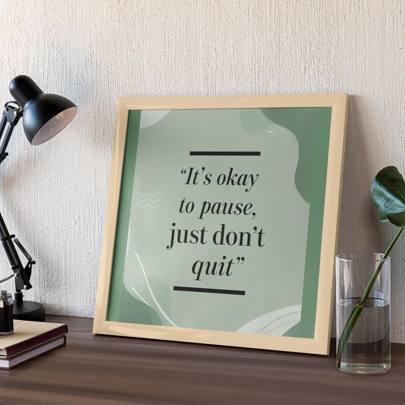 May include: A framed print with the quote "It's okay to pause, just don't quit" in black text on a green background. The frame is light beige, and the print is displayed on a wooden surface with a black desk lamp and a glass vase.