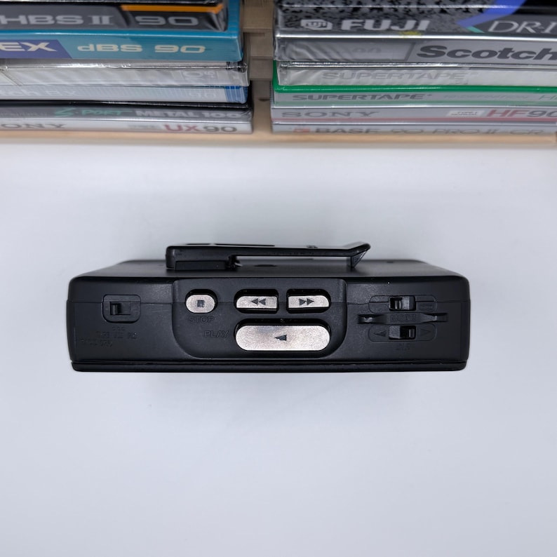 May include: A black portable cassette player with silver buttons for play, rewind, and fast forward. The device has a clip on the top and is positioned below a shelf holding several cassette tapes.