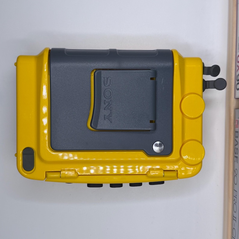 K&ouml;nnte beinhalten: Ein gelb-grauer Sony Walkman-Kassettenrekorder. Der Player hat eine rechteckige Form mit abgerundeten Kanten und einer Klappt&uuml;r. Das Sony-Logo ist oben sichtbar. Der Player hat zwei runde Kn&ouml;pfe und ein kleines schwarzes Bedienfeld.