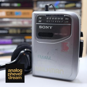 Peut inclure: Un lecteur de cassettes portable Sony Walkman argent&eacute; avec un casque audio attach&eacute;. L'appareil dispose de capacit&eacute;s radio FM/AM et pr&eacute;sente le logo "Walkman". Le texte "analog phever dream" est dans le coin inf&eacute;rieur gauche.