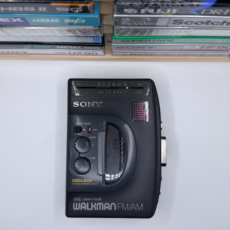 May include: Black Sony Walkman FM/AM radio with Mega Bass and Auto Reverse features. The device has a digital display and control buttons. The model number is WM-FX38. A collection of cassette tapes is visible in the background.