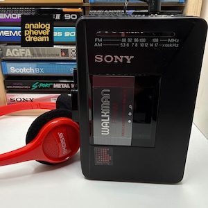 May include: A black Sony Walkman FM/AM radio cassette player with a silver center panel and a red pair of Koss headphones. The device features a frequency display for FM and AM bands, and the text Walkman FM/AM Radio Cassette Player WM-AF23.