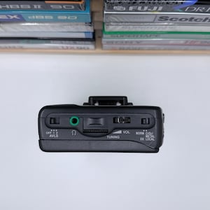 May include: Black portable cassette player with a silver volume dial and a green audio jack. The player has buttons labeled "OFF 1 2 AVLS", "TUNING", and "NORM CrO2/METAL DX LOCAL". Cassette tapes are visible in the background.
