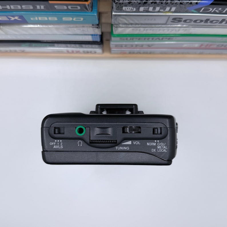 May include: Black portable cassette player with a silver volume dial and a green audio jack. The player has buttons labeled "OFF 1 2 AVLS", "TUNING", and "NORM CrO2/METAL DX LOCAL". Cassette tapes are visible in the background.