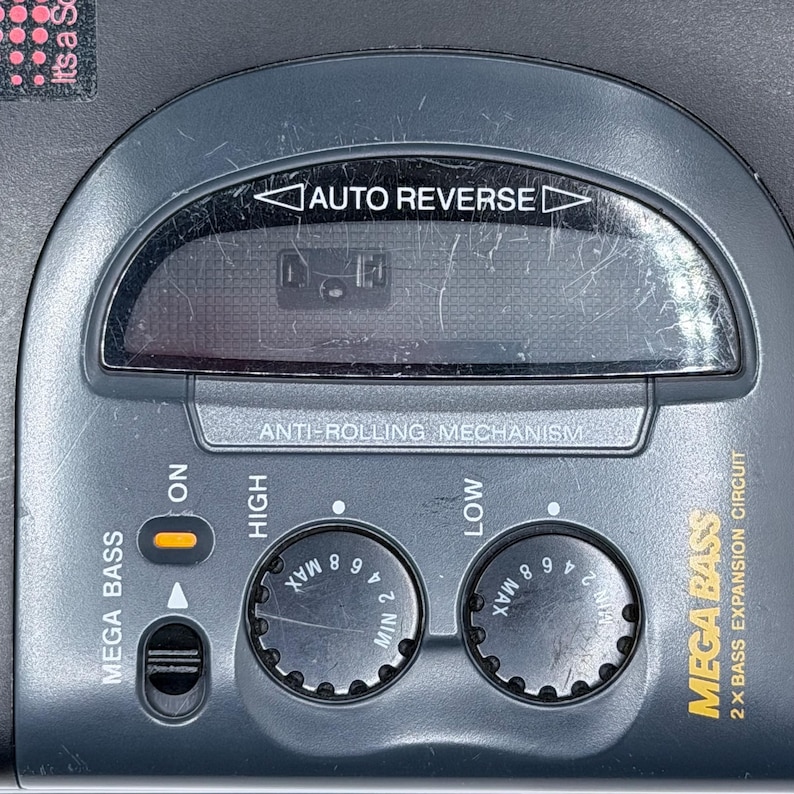 May include: Close-up of a gray portable cassette player with the text "MEGA BASS" and "AUTO REVERSE". It has dials for bass and volume control, and a display window. The player also has an "ON" button.