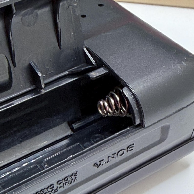 May include: Close-up of a black Sony WM-FX10 radio cassette player's battery compartment. The compartment is open, revealing a silver spring and the interior details. The Sony logo is visible on the side.