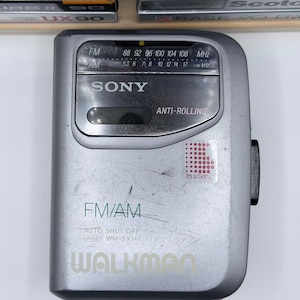 Peut inclure: Un lecteur de cassettes portable Sony Walkman argent&eacute; avec radio FM/AM. L'appareil comporte un cadran de r&eacute;glage, un &eacute;cran et le texte "WALKMAN" en bas. La radio porte l'inscription "It's a Sony" sur un autocollant.