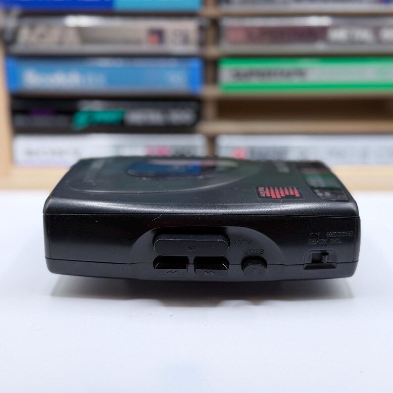 May include: A black portable CD player with buttons for play, stop, and other functions. The device has a digital display and a headphone jack. Cassette tapes are visible in the background.