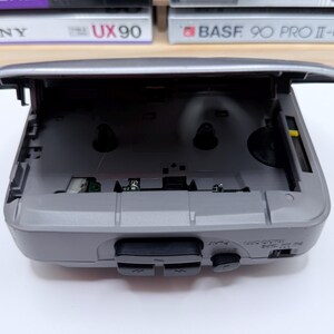 Peut inclure: Un lecteur de cassettes portable gris avec le dessus ouvert, r&eacute;v&eacute;lant le compartiment &agrave; cassettes. Le lecteur a des boutons noirs pour la lecture, l'arr&ecirc;t et d'autres fonctions. Le dessus du lecteur est ouvert, et la cassette n'est pas ins&eacute;r&eacute;e. Le lecteur est sur une surface blanche.
