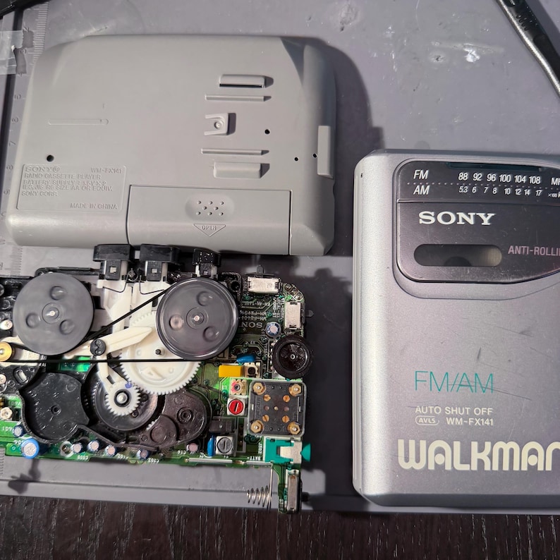 Peut inclure: Radio Sony Walkman FM/AM argent&eacute; d&eacute;mont&eacute;, avec le couvercle arri&egrave;re ouvert, r&eacute;v&eacute;lant les composants internes. La radio a un cadran de fr&eacute;quences et le texte "WALKMAN" est visible. Le bo&icirc;tier en plastique gris est ouvert, montrant le circuit imprim&eacute; et les pi&egrave;ces m&eacute;caniques.