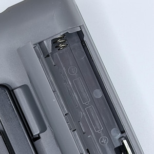 Peut inclure: Gros plan sur un compartiment &agrave; piles en plastique gris, probablement d'une t&eacute;l&eacute;commande ou d'un appareil similaire. Le compartiment est ouvert, r&eacute;v&eacute;lant les contacts &agrave; ressort et les emplacements pour les piles. Le design est simple et fonctionnel.