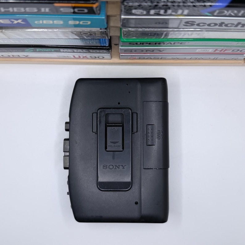May include: A black Sony Walkman cassette player with a belt clip. The player has buttons on the side and a release latch. Cassette tapes are visible in the background.