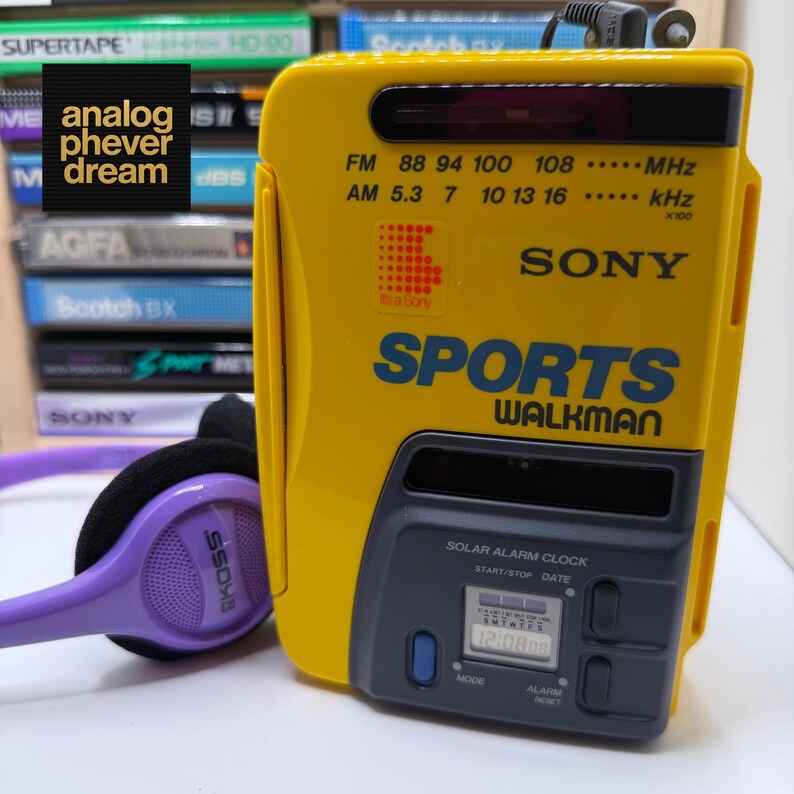 K&ouml;nnte beinhalten: Ein gelber Sony Sports Walkman mit blauer Schrift und digitaler Uhranzeige. Der tragbare Kassettenrekorder hat einen schwarzen oberen und einen grauen unteren Bereich. Ein lila Koss-Kopfh&ouml;rer liegt daneben.