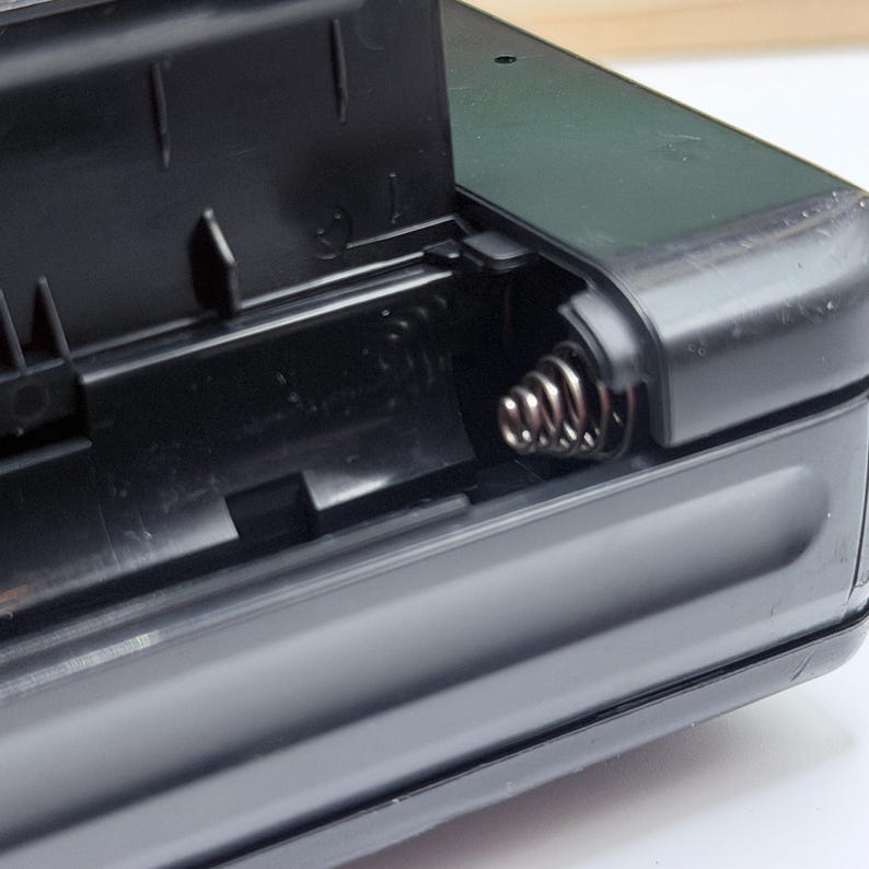 May include: Close-up of a black plastic battery compartment with the lid open. A silver spring is visible inside the compartment. The compartment is part of a larger black electronic device.