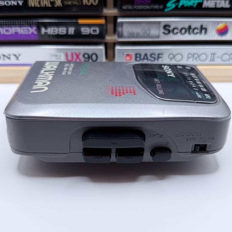Peut inclure: Un lecteur de cassettes Sony Walkman gris vintage. L'appareil a une bordure gris fonc&eacute;, des boutons noirs et le mot "WALKMAN" imprim&eacute; sur le dessus. Le lecteur est pr&eacute;sent&eacute; avec une collection de cassettes en arri&egrave;re-plan.