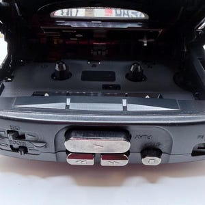 May include: A black portable cassette player with the top open, revealing the tape compartment. Silver buttons for play, stop, rewind, and fast forward are visible. The device has a mode and direction button.