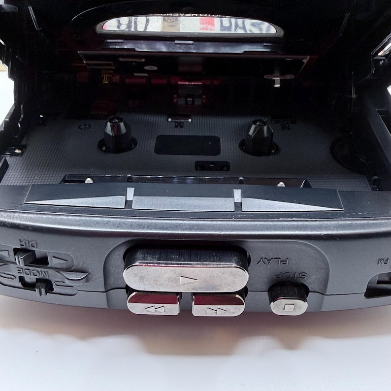 May include: A black portable cassette player with the top open, revealing the tape compartment. Silver buttons for play, stop, rewind, and fast forward are visible. The device has a mode and direction button.