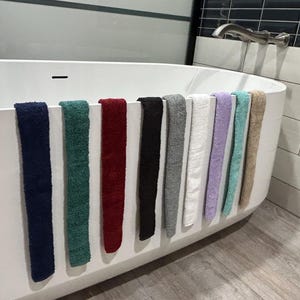 May include: A collection of colorful bath towels in various shades, including navy, green, red, black, gray, white, purple, teal, and beige, hanging on the edge of a white bathtub. The towels are soft and fluffy.